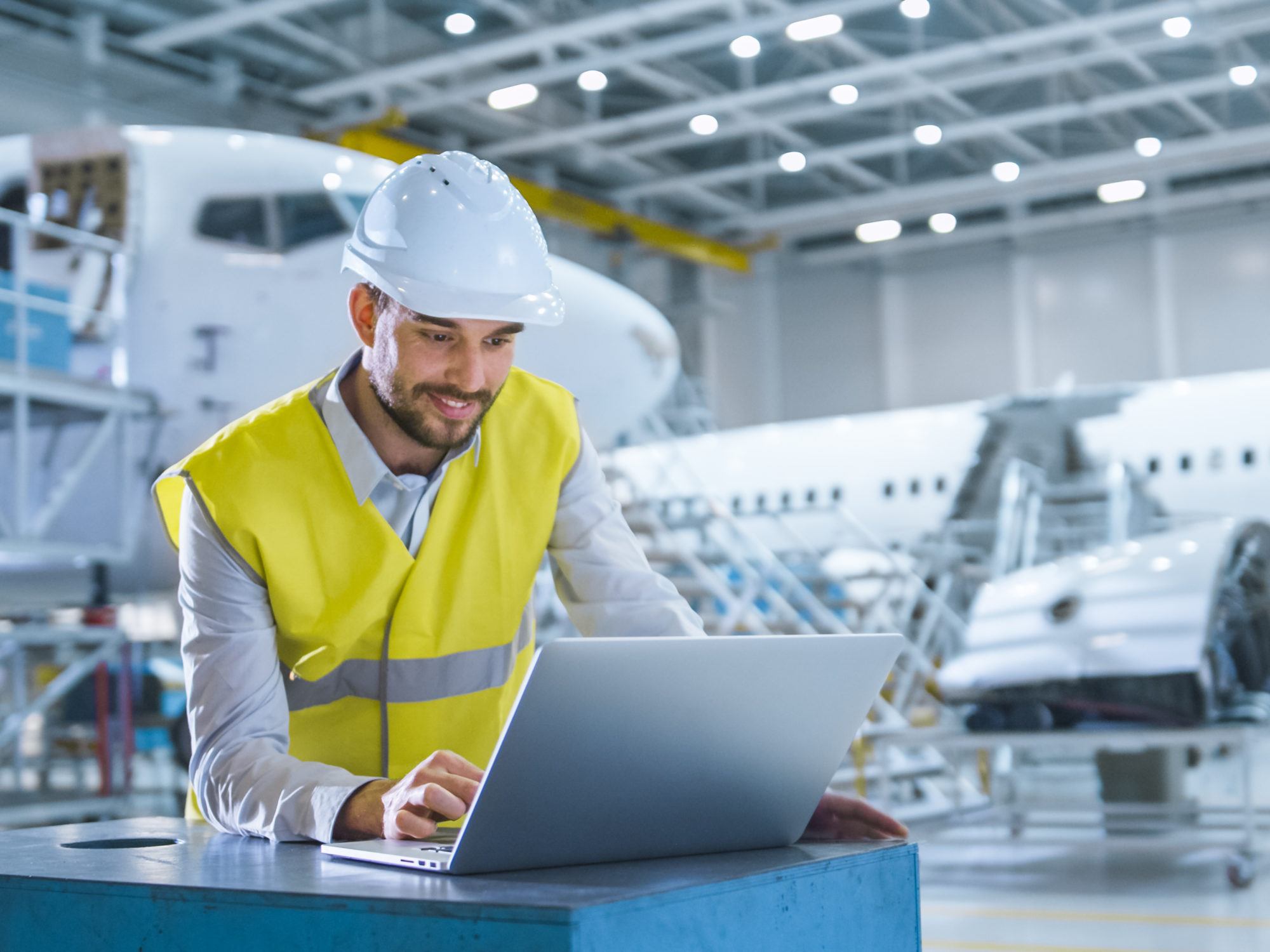 engineer in safety vest working on his laptop in front of aircraft maintenance hangar