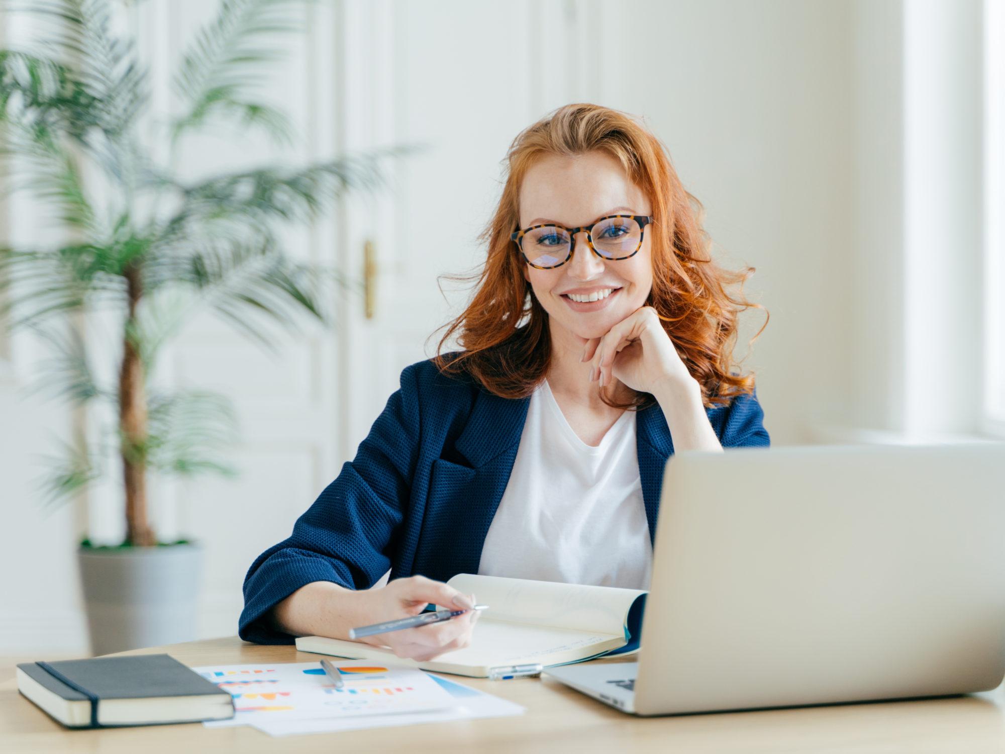 Photo of successful ginger female freelancer has remote work, watches webinar online on laptop computer, writes down information in notepad, smiles happily poses at workplace dressed in formal clothes