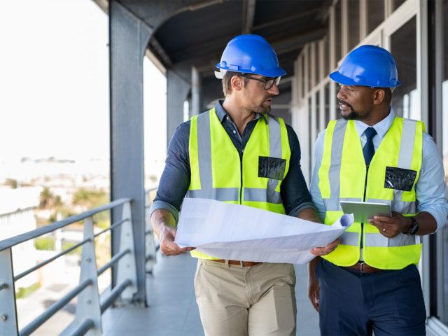 workers in hardhats walking and discussing the sheet one of them is holding