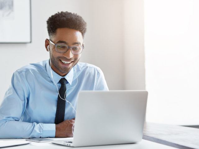 Aufnahme eines fröhlichen erfolgreichen jungen Managers afroamerikanischer Abstammung mit Dreitagebart in einem Büro, der vor einem geöffneten Laptop sitzt und Ohrhörer trägt, während eine Videokonferenz mit Geschäftspartnern läuft.