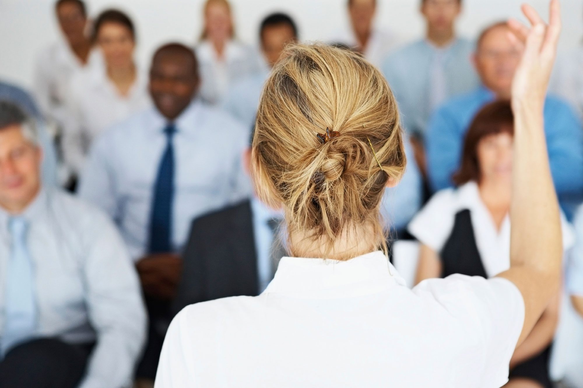 View of female back of head raising hand and speaking in front of crowd