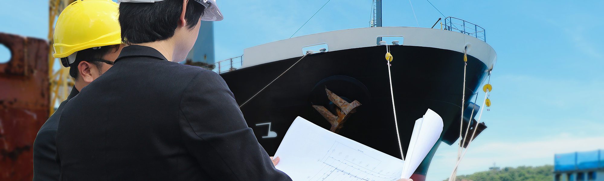 Two engineers reading a blueprint by front cargo ship or shipbuilding in shipyard background